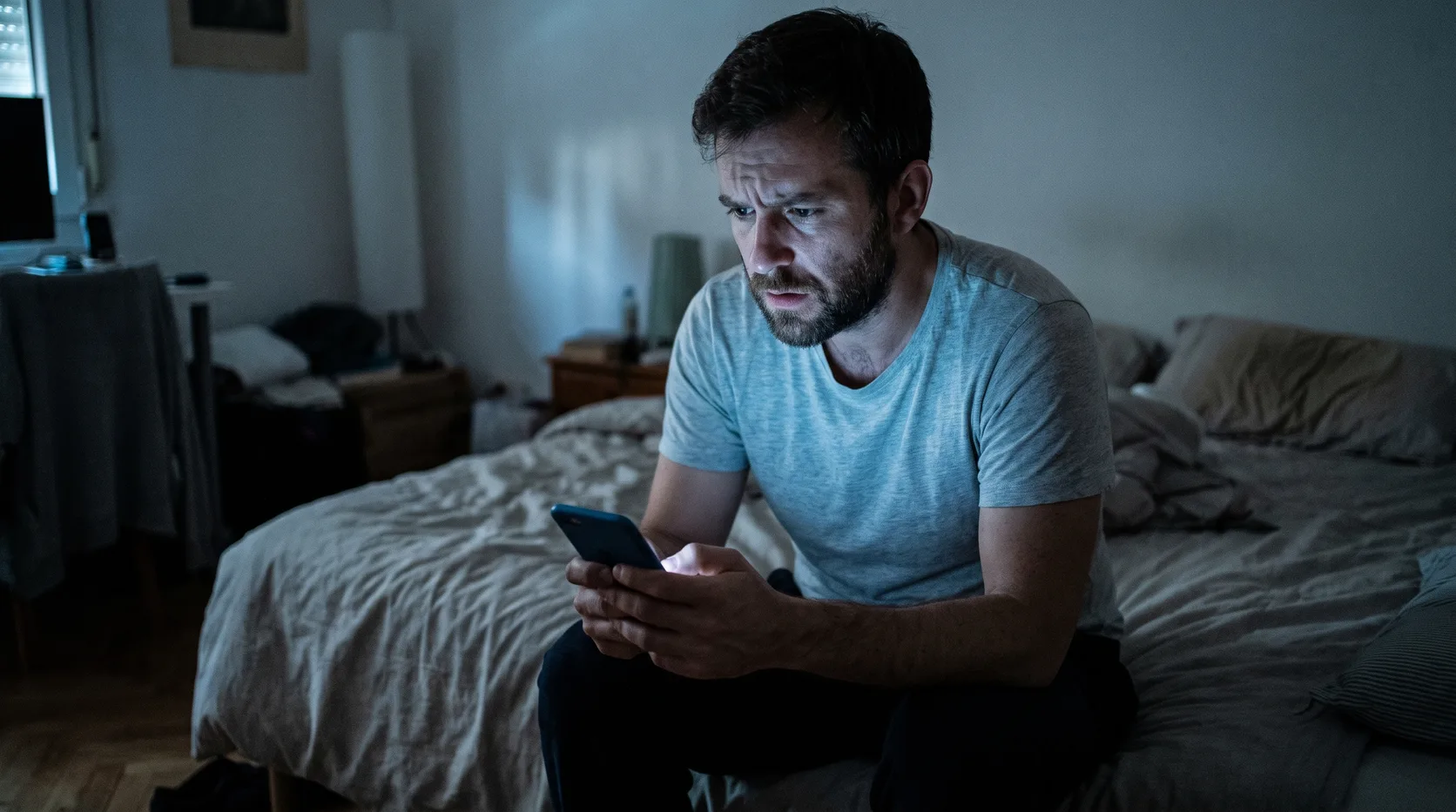 A man sitting on the edge of a bed at night, his face lit by the glow of a phone screen