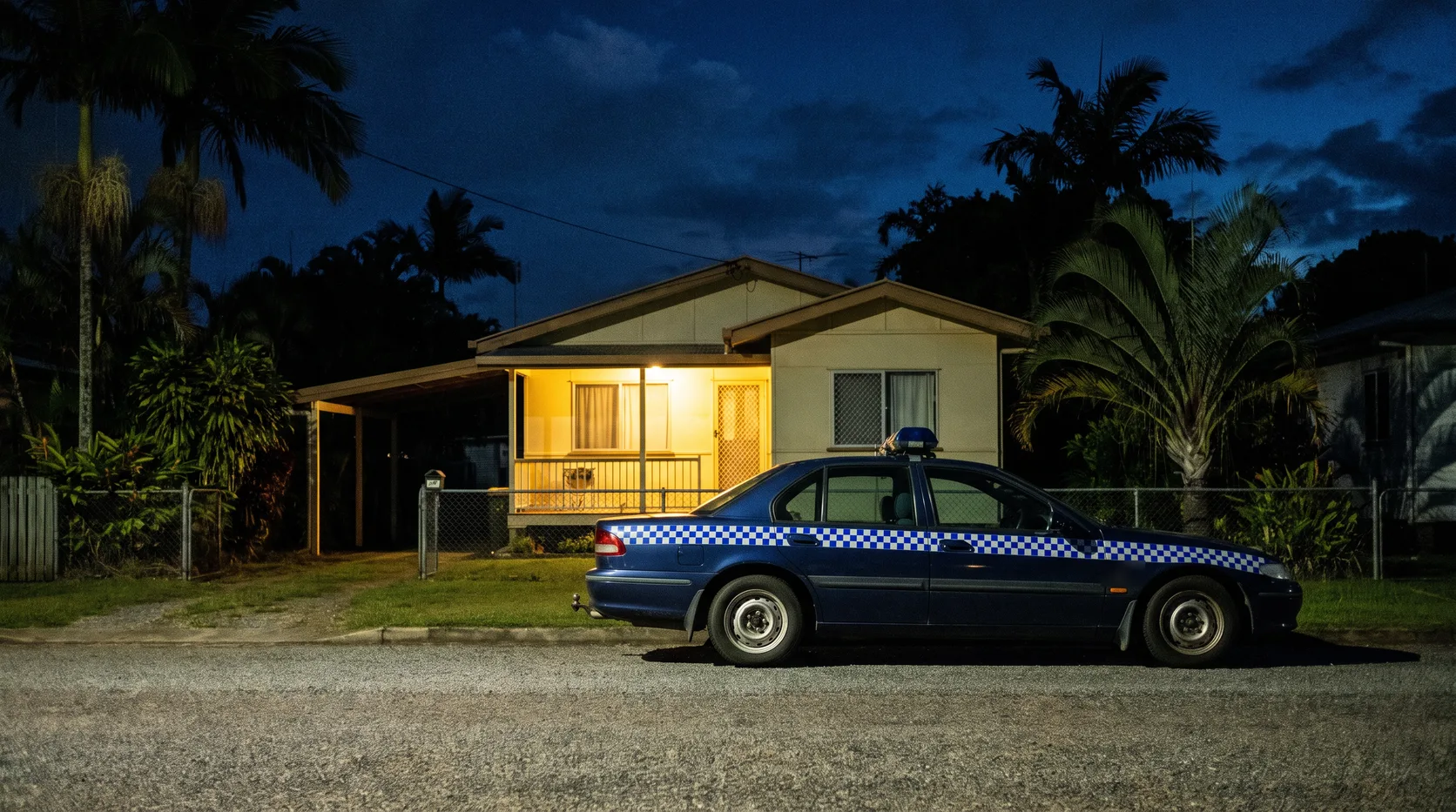 A police car parked at night outside a fibro house on a quiet suburban street