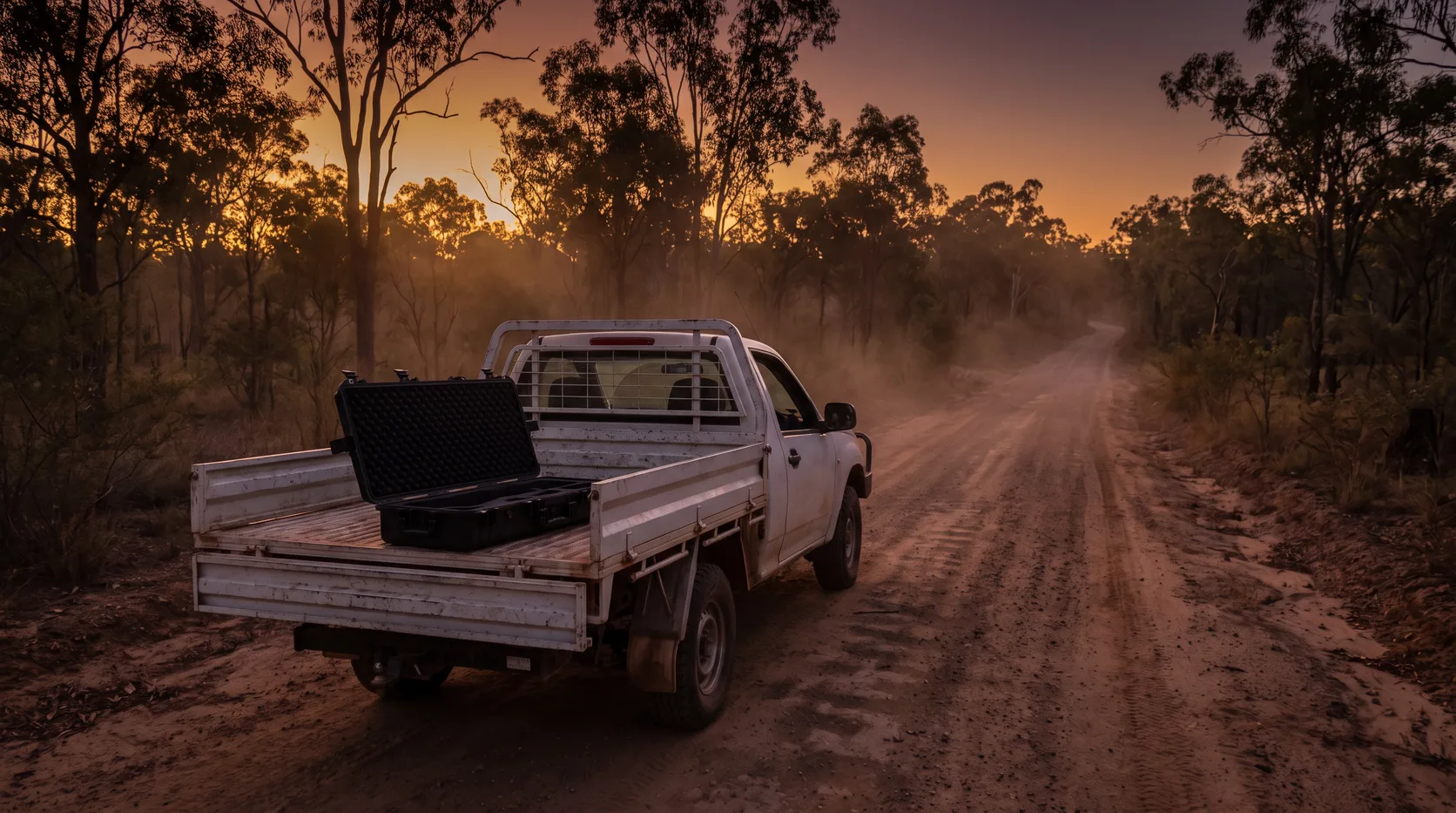 The open boot of a ute parked on a dirt track in the bush at dusk, with an empty hard case inside
